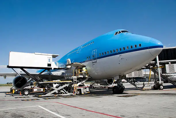 Boeing 747 aircraft being catered and loaded on the platform. The catering truck highloader is parked in front of one of the doors. Image taken under a blue sky with a side front view, cockpit and nose of the airplane to the right.
