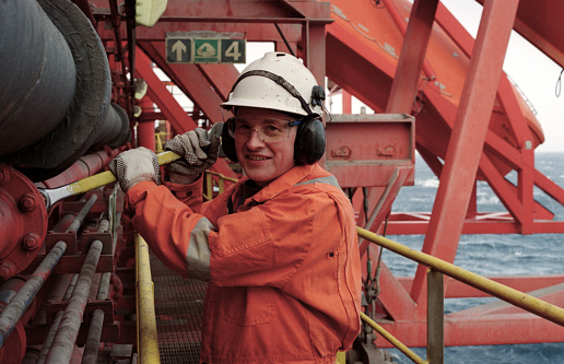 Roughneck, engineer or male technician working with a large spanner on a pipe outdoors on a north sea oil drilling rig. Wearing safety equipment, coveralls, hard hat and gloves. Work, employment, occupation and oil industry are common themes.