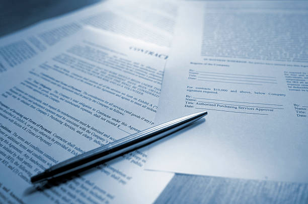 Contract A blue tinted image of silver ball point pen lying on scattered legal contract documents on a wooden table, ready to sign to complete the deal. This conceptual image is shot with a shallow depth of field, and all paperwork is generic. Part of a contract series of images.  legal document stock pictures, royalty-free photos & images