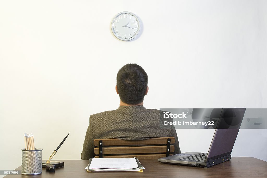 A man sitting backing against a desk looking up at a clock Man in his office waiting for the day to end Wasting Time Stock Photo