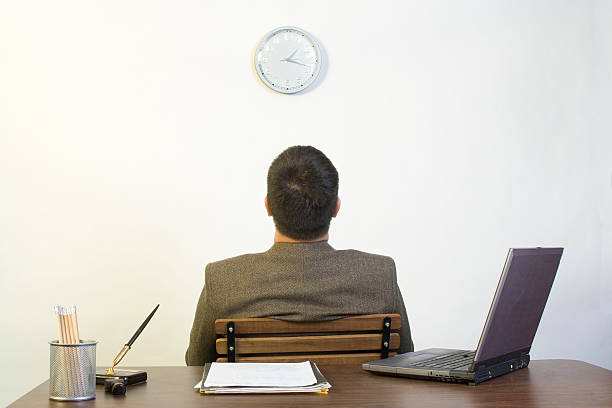 A man sitting backing against a desk looking up at a clock Man in his office waiting for the day to end wasting time photos stock pictures, royalty-free photos & images