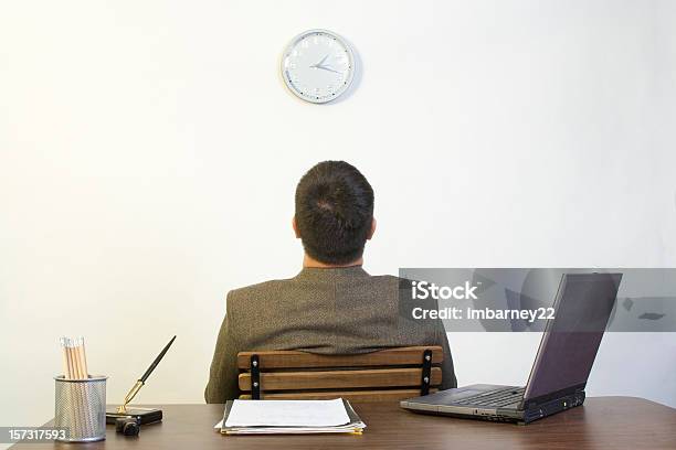 A Man Sitting Backing Against A Desk Looking Up At A Clock Stock Photo - Download Image Now