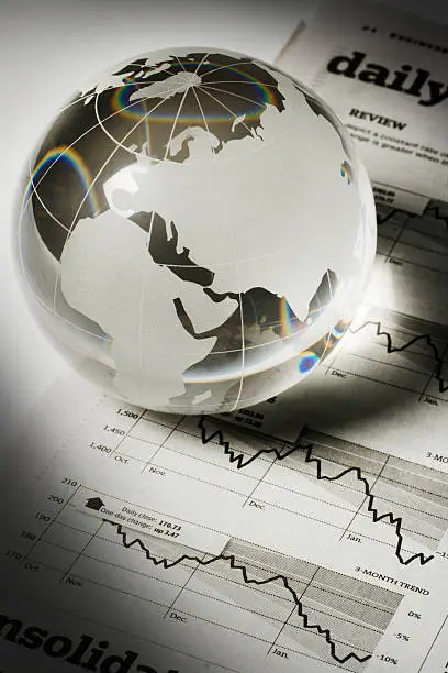 A glass globe paperweight resting on newspaper financial page charts and graphs, which indicate downward global business, investment, and finance trends. The earth sphere casts a dark, gloomy shadow, implying forecasting economic recession and fortune telling stock market trouble. Europe, Africa, and Asia are visible, for concepts of world economy.