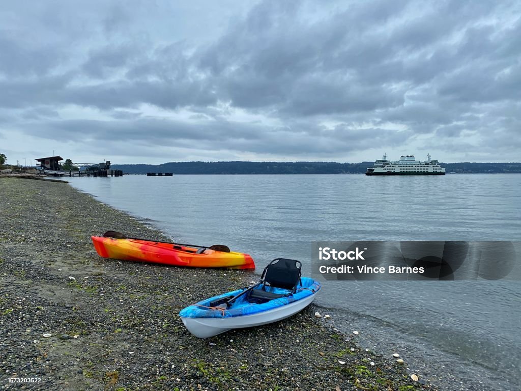 Kayaks in Mukilteo, Washington With the Mukilteo-Whidbey Island Ferry in the background, two kayaks rest on the rocky shore of Edgewater Park, in Mukilteo, Washington. Aquatic Sport Stock Photo
