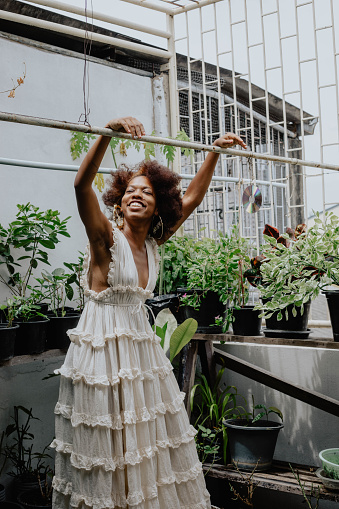 Woman laughing with her arms above her head standing in her rooftop garden wearing a flowy  white dress