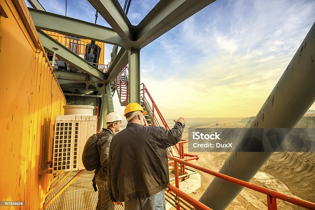 Heavy mining industry workers Two coal mine engineers with protective helmets standing on a huge drill machine, chatting and watching at the digging site. Beautiful and colorful sky in the background. Rear view. Mining - Natural Resources Stock Photo