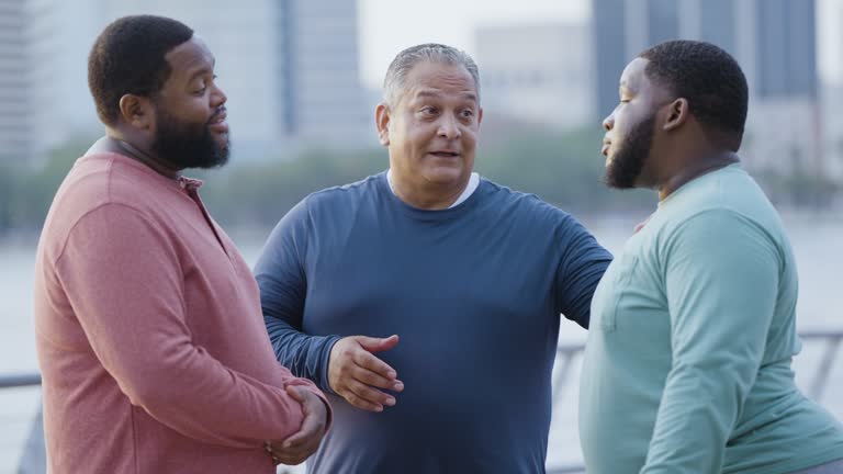 Three multiracial heavyset men talk on city waterfront