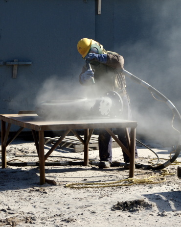 Man sand blasting a section of piping that belongs to a fishing boat. He is wearing protective gear to do his job.