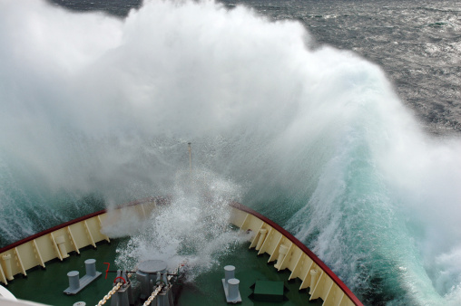 A ship slamming its bow into a large oncoming wave, making it break over the bow.