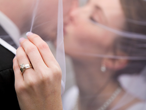 A close up of a bride's wedding ring as the couple kisses in the background.