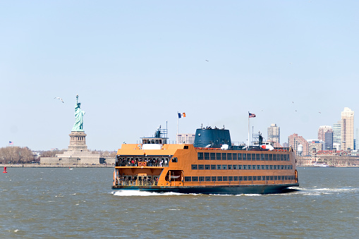 Staten Island ferry coming from Manhattan and passing by the Statue of Liberty. The Statue of Liberty and the skyscrapers of lower manhattan can be seen in the background under a clear sky.