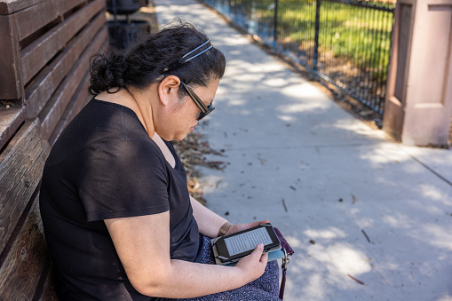 High quality stock photo of an Asian American woman reading an e-reader in the park.