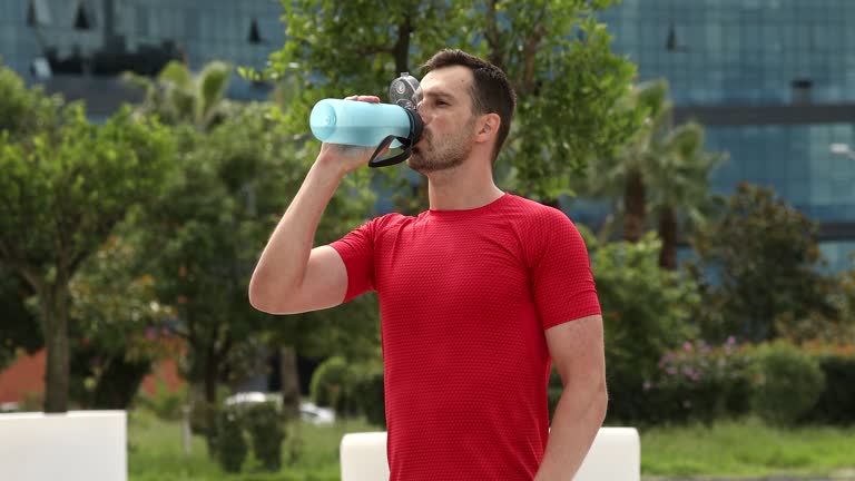 Young muscular man wearing red sports shirt walking in park and breathing hard, drinking water from bottle. Concept of fitness.