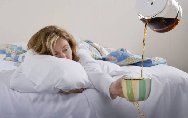 A woman in bed holding an overflowing coffee mug in her hand Exhausted woman in bed wanting more coffee. caffeine stock pictures, royalty-free photos & images