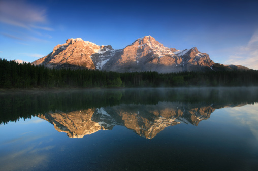 Three mountain peaks reflected in a lake. Beautiful Wedge Pond in Kananaskis Country reflecting Mount Lorette and Mount Kidd. Nobody is in the image. Beautiful serene summer scenic in Alberta, Canada. This is a photographer's dream with mist coming off the pond and a beautiful sky as well as soft morning light on the pond. There is no wind which makes for a perfect reflection. 
