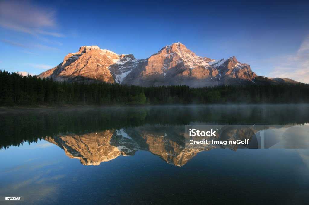 Canmore Canadian Rockies Mountain Scenic Three mountain peaks reflected in a lake. Beautiful Wedge Pond in Kananaskis Country reflecting Mount Lorette and Mount Kidd. Nobody is in the image. Beautiful serene summer scenic in Alberta, Canada. This is a photographer's dream with mist coming off the pond and a beautiful sky as well as soft morning light on the pond. There is no wind which makes for a perfect reflection.  Kananaskis Country Stock Photo