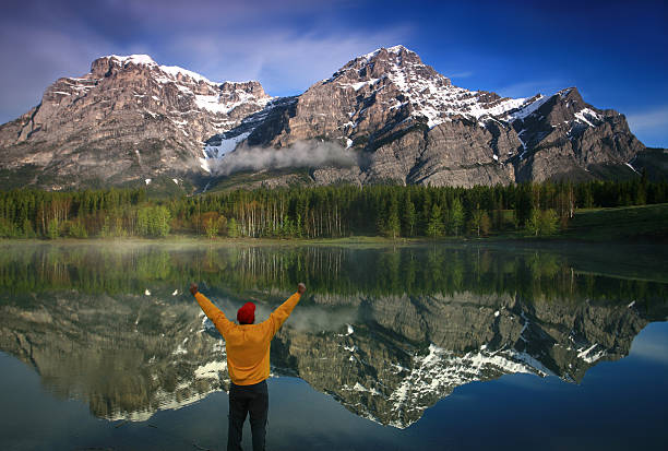 Happy Caucasian Middle Aged Man in the Mountains A happy man in a beautiful mountain scene. Image location is in Kananaskis Country Alberta, Canada. The model is a middle aged Caucasian man who is unrecognizable. Back view. Beautiful reflection of Mount Lorette in the mist-covered lake. Man is wearing bright colours. Additional themes in the image are joy, happiness, beauty, nature, hiking, naturalist, nature lover, arms raised, content, one person, men, alone, and natural beauty.  kananaskis country stock pictures, royalty-free photos & images