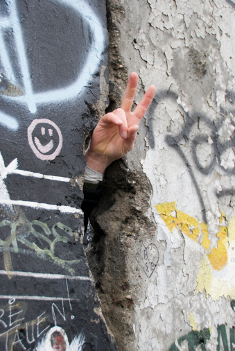 Close up of a hand through a crack in the Berlin Wall, making the PEACE symbol. On the wall graffiti and a smile.