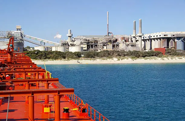 Large industrial plant, alumina refinery on beach in Western Australia. Orange-red coating of bulk carrier in foreground.