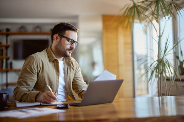 istock Young man working on reports and laptop at home office. 1573363467