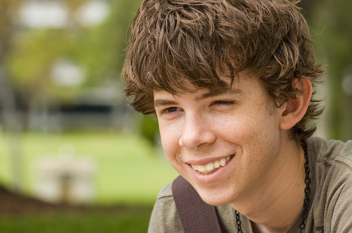 A teenage male student has a big smile in this closeup head and shoulders outdoors portrait. Shallow depth of field.