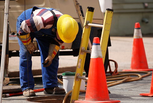 A construction worker looks down a man hole.  He is bent over wearing safety gear, hard hat and gloves.  There is a ladder down and safety cones for traffic control.