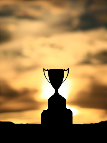 A silhouette of a silver trophy cup against dramatic sunset. Themes of the image include competition, winning, success, achievement, winner, champion, championship, and prizes. Nobody is in the image. Trophy is an authentic silver cup that was presented to a champion of a baseball tournament. Vertical colour image. 