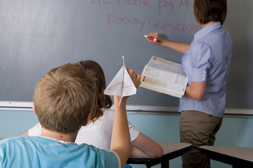 Student aiming paper plane at teacher writing on blackboard