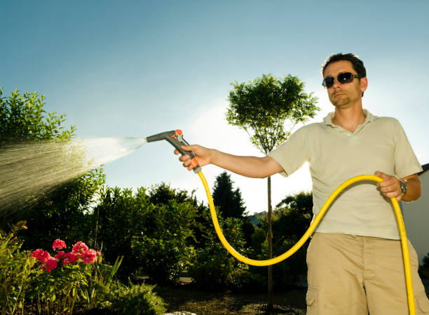 Defocus closeup water garden. Female hand holding blue automatic sprinkler sprays water over brown blurred background. Gardener watering the lawn in the garden. Sunflare. Out of focus.