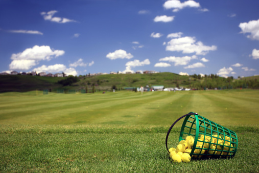 A driving range with practice balls and bucket. Yellow practice golf balls are common at driving range facilities throughout North America and the world. Driving ranges often sell plastic buckets of small, medium, and large sizes. Themes in this image include golf industry, turf grass, practice balls, golf balls, driving range buckets, practice facilities, golf resorts, learning, lessons, golf lessons, and fairway. Nobody is in the image. 