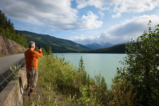 Senior man taking photo of scenic turquoise lake and mountainous landscape at a viewpoint at the side of a road.  British Columbia, Canada.