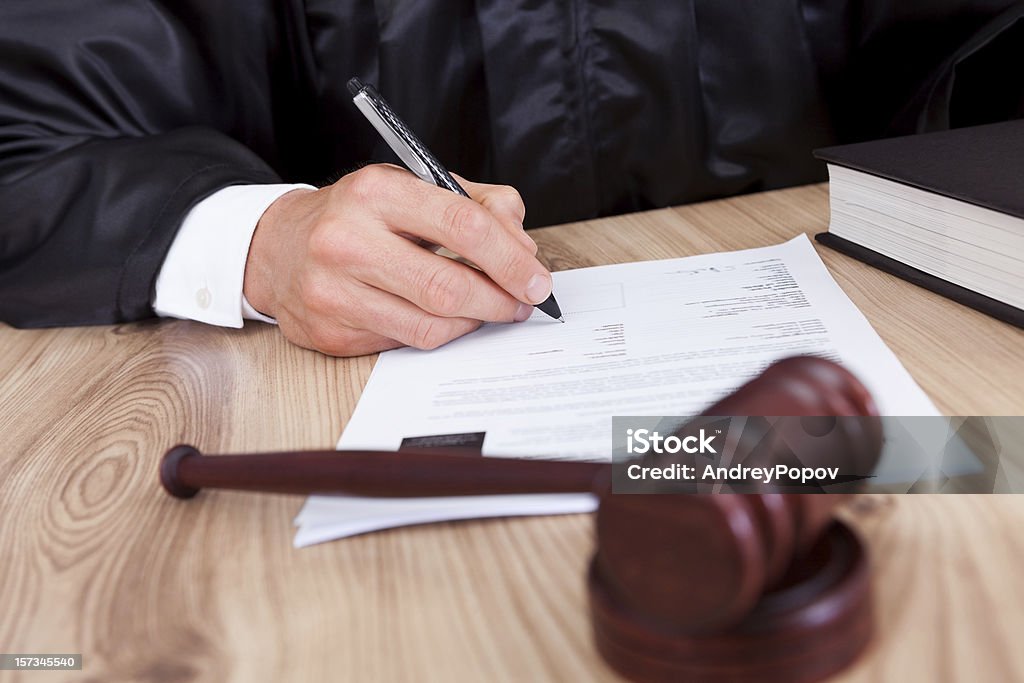 Male judge signing papers with a gavel in front of him Male Judge Writing On Paper In Courtroom Judge - Law Stock Photo