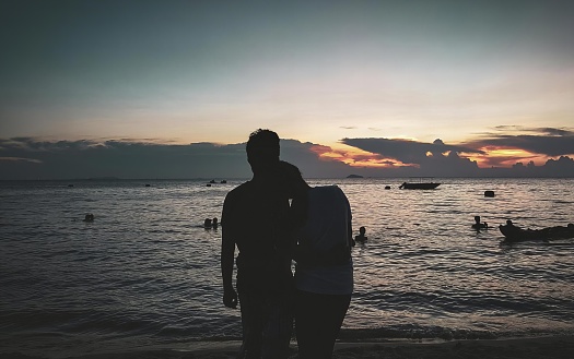 a photography of a couple standing on the beach at sunset, there are two people standing on the beach watching the sun set.