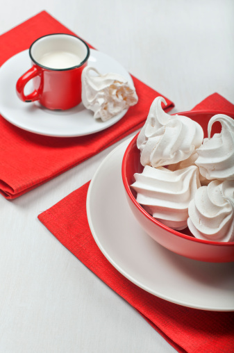 View of a red bowl with white meringues on a red textile napkin. In the distance is a red cup with milk and a broken piece of meringue on a plate.