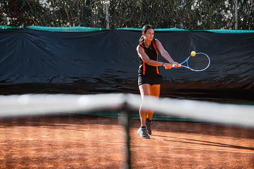 Woman in black outfit training under a harsh sun on a clay court.