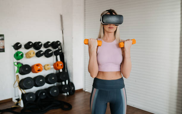istock Focused young blonde woman in sportswear training with dumbbells using virtual reality headset as a trainer stands at gym against shelves with weights. American girl with great body shape at exercise. 1573535270