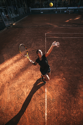Young woman in black outfit serves ball during a match in daylight under a harsh sun.