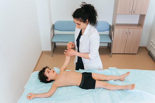 Young orthopaedic specialist examining little curly boy holding his hand. Preschooler hispanic boy laying on medical bench at hospital receiving therapeutic massage. Medicine, healthcare.