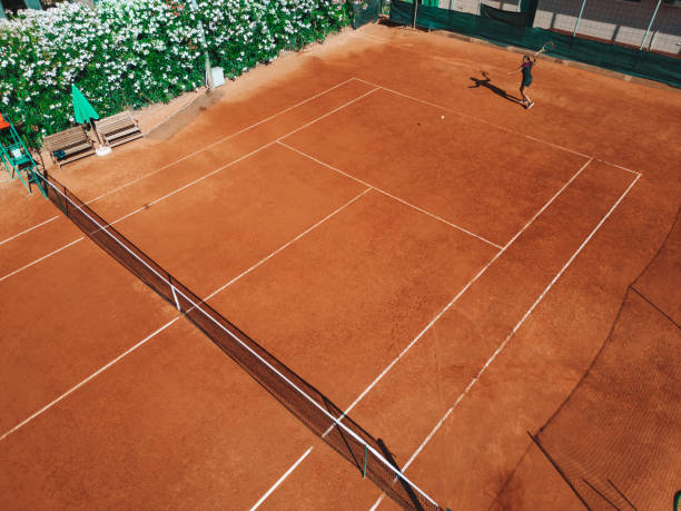 Woman in black outfit training under a harsh sun on a clay court seen from a drone point of view.