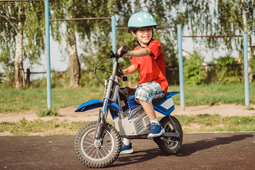 A little boy in a protective helmet rides a children's electric motorcycle outdoors in a park on a sunny summer day
