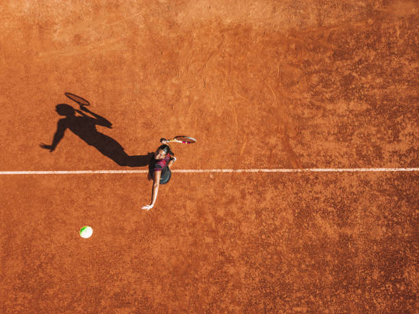 Professional tennis player serves - aerial point of view Drone point of view of a young woman in black outfit serves ball during a match in daylight under a harsh sun. clay court stock pictures, royalty-free photos & images