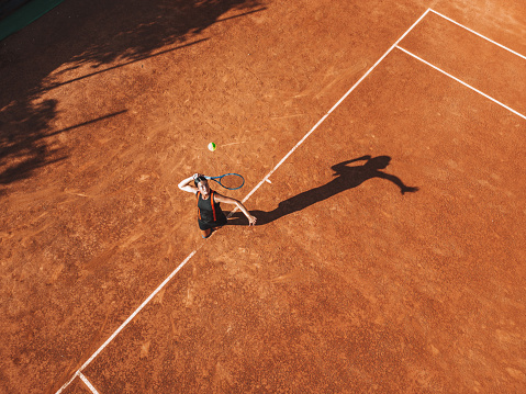 Drone point of view of a young woman in black outfit serves ball during a match in daylight under a harsh sun.
