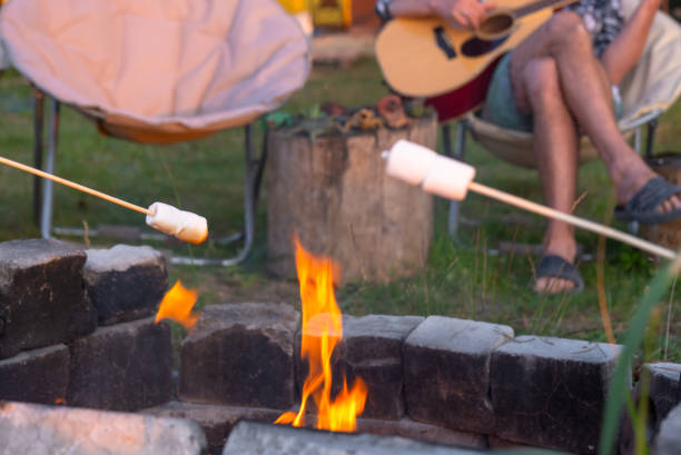 une fille en chemise à carreaux fait rôtir des guimauves sur un feu dans la cour de la maison, le père joue de la guitare. soirée familiale près du feu de camp, pique-en plein air dans des fauteuils touristiques - historical reenactment fun heat recreational pursuit photos et images de collection