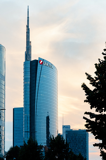 Milan, Lombardy, Italy- June 27, 2023; At dusk in a neighborhood with office buildings in Milan-Italy, the sun sets behind a large futuristic office building. The building has a large glass facade reflecting the sunset.