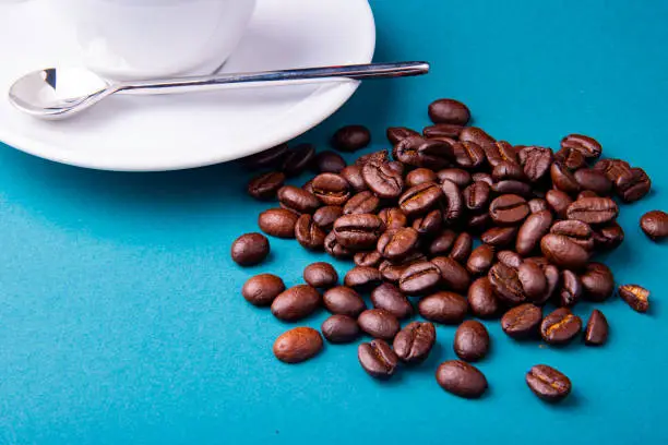 White espresso cup and saucer with brown coffee on a teal blue surface with beans and spoon.