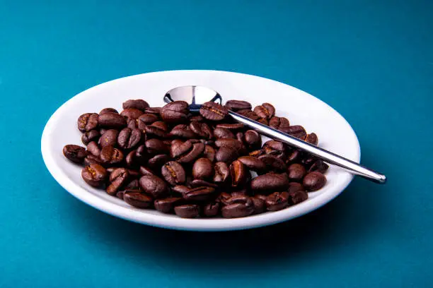 White espresso cup and saucer with brown coffee on a teal blue surface with beans and spoon.