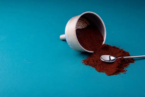 Aluminium filter with brown coffee beans on a teal blue surface with ground beans.