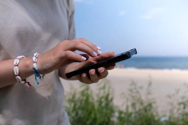 istock Female hand with shell bracelets holding smartphone. Unrecognizable woman using mobile phone on the beach seacoast. Holiday vacation advertisement. Social media 1573721594