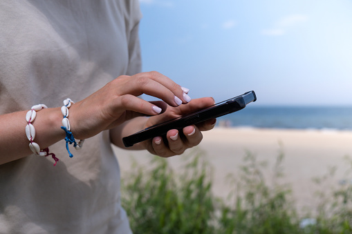 Female hand with shell bracelets holding smartphone. Unrecognizable woman using mobile phone on the beach seacoast. Holiday vacation advertisement concept. Social media