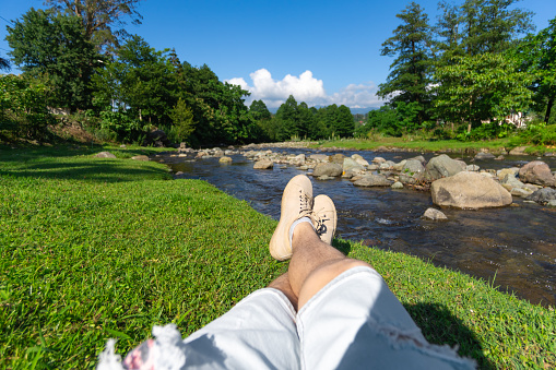 Close-up of the legs of a man in denim shorts and sports shoes sitting on the green grass by a mountain river on a sunny day. Focus on the legs